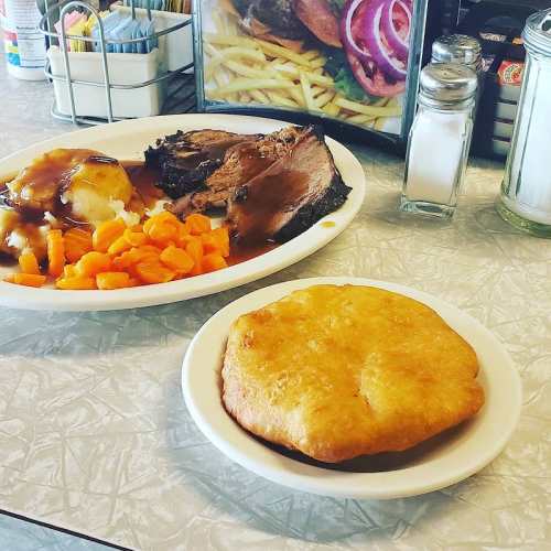 A plate of sliced roast beef with gravy, mashed potatoes, carrots, and a side of fried bread on a diner table.