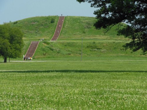 A grassy field with a large earthen mound in the background, featuring a sloped path and people walking on it.