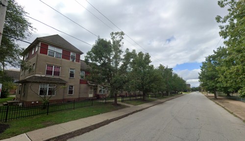 A quiet street lined with trees and a multi-story building on one side under a cloudy sky.