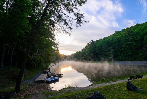 A serene lake at sunrise, surrounded by trees, with mist rising from the water and boats docked nearby.