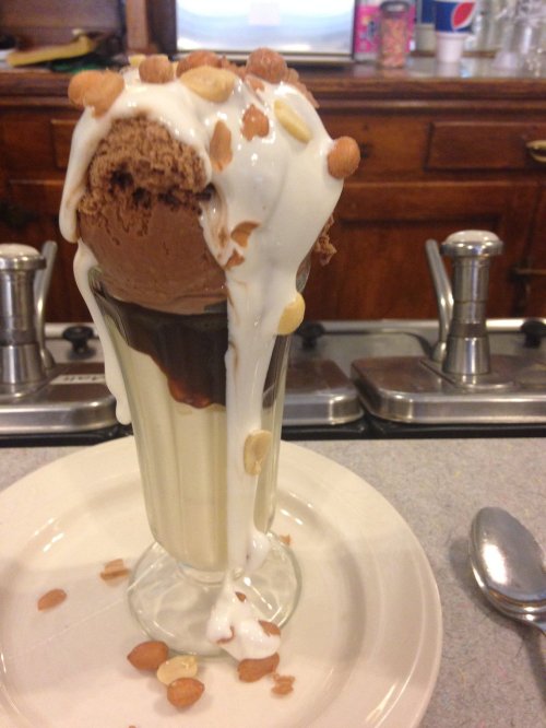 A tall glass dessert with layers of chocolate and vanilla ice cream, topped with whipped cream and nuts, dripping slightly.