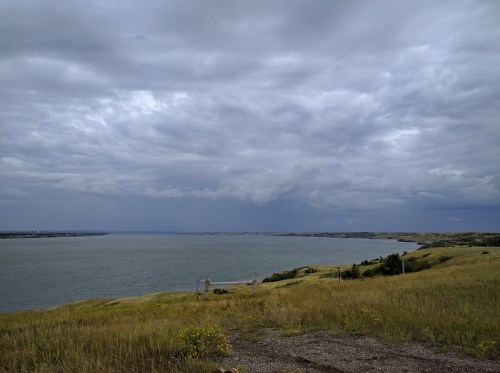 A scenic view of a calm lake surrounded by grassy hills under a cloudy sky.