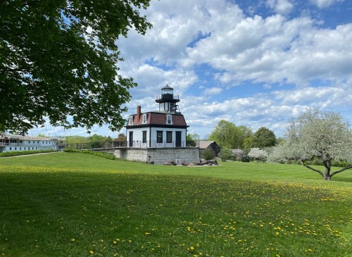 A historic lighthouse stands on a grassy hill, surrounded by trees and blooming flowers under a partly cloudy sky.