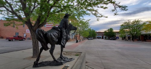 A bronze statue of a cowboy on a horse, surrounded by trees and a quiet street in a small town.