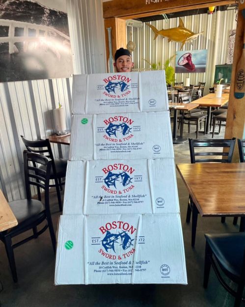 A person stands behind stacked boxes labeled "Boston Sword & Tuna" in a casual dining setting.