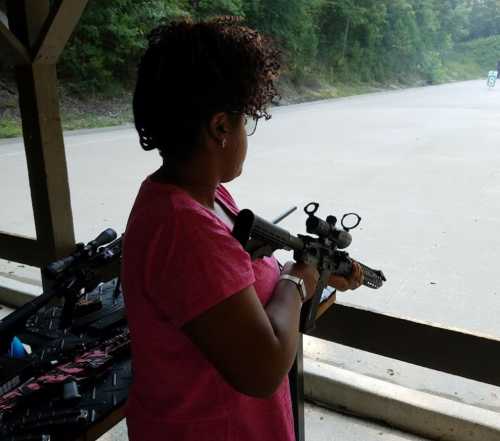 A woman with curly hair aims a rifle at a shooting range, with firearms visible on a table behind her.