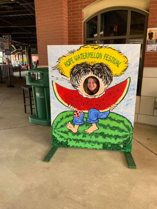 A person peeks through a watermelon-themed cutout at the Hope Watermelon Festival, smiling and posing playfully.