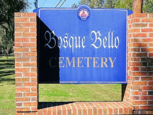 Sign for Bosque Bello Cemetery, featuring blue background and brick structure, surrounded by green grass and trees.
