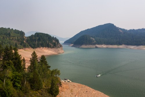 A serene lake surrounded by forested hills, with a small boat gliding across the water under a hazy sky.
