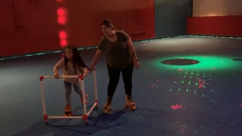 A woman assists a young girl on roller skates using a support frame in a colorful skating rink.
