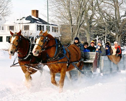 A horse-drawn sleigh carries a group of people through a snowy landscape, with a house in the background.