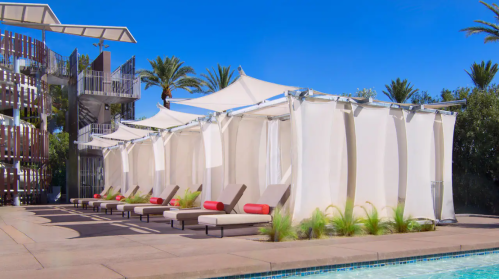 A poolside area with lounge chairs and white cabanas under a clear blue sky, surrounded by palm trees.