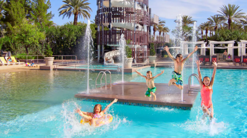 Four children joyfully jumping into a pool with a water slide in the background, surrounded by palm trees.