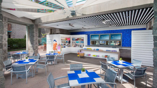 Bright outdoor café with blue tables, chairs, and a snack counter, surrounded by a modern architectural design.