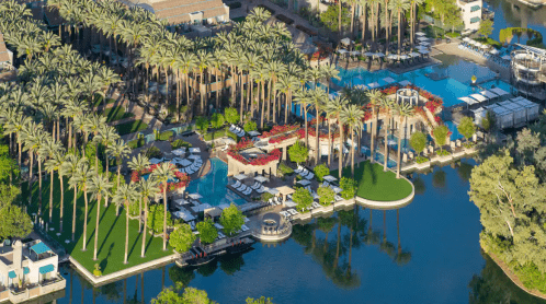 Aerial view of a luxurious resort with palm trees, pools, and a serene lake surrounded by lush greenery.