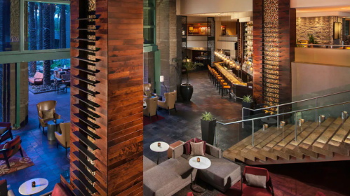 Modern hotel lobby with wooden accents, cozy seating areas, and a bar in a spacious, well-lit environment.