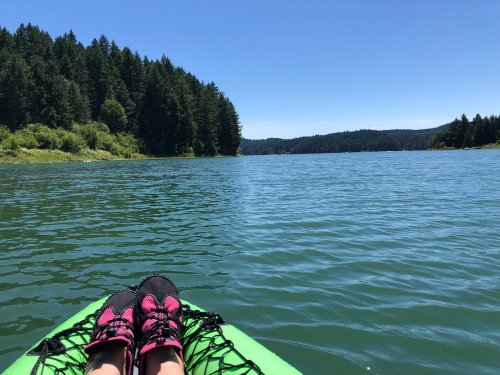 A pair of feet in black and pink shoes resting on a green kayak, with a serene lake and forested hills in the background.