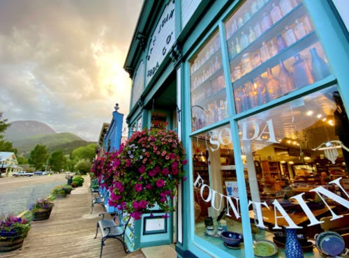 A charming soda fountain shop with colorful flowers and vintage bottles, set against a mountain backdrop at sunset.