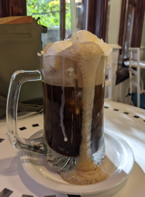 A frosty mug of root beer floats over with foam and ice cream, sitting on a white table.