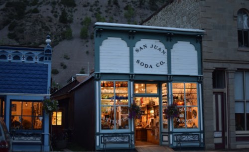 A vintage soda shop with large windows, displaying colorful bottles, set against a mountain backdrop at dusk.