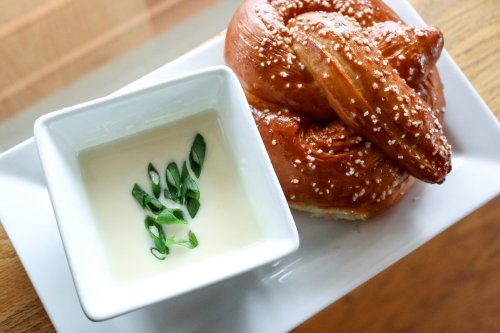 A soft pretzel with coarse salt served alongside a small bowl of creamy dip garnished with green onions.