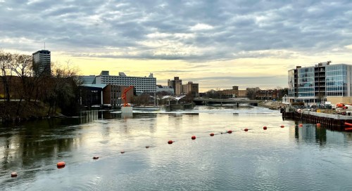 A river scene with buildings, a sculpture, and a cloudy sky reflecting on the water's surface.