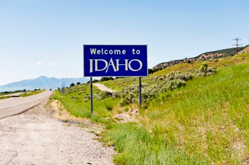 A blue welcome sign for Idaho beside a highway, with green grass and mountains in the background under a clear sky.