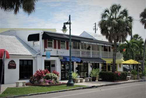 A charming street view of a two-story building with shops, palm trees, and outdoor seating under yellow umbrellas.