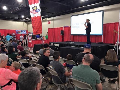 A speaker stands on stage addressing an audience at a convention, with red curtains and banners in the background.