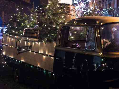 A vintage truck decorated with Christmas lights and filled with small trees, set against a festive backdrop.