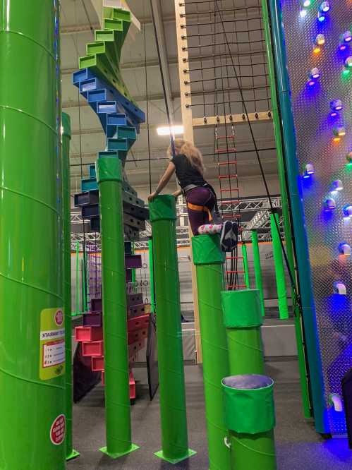 A person climbs on green vertical structures in an indoor obstacle course, surrounded by colorful climbing elements.