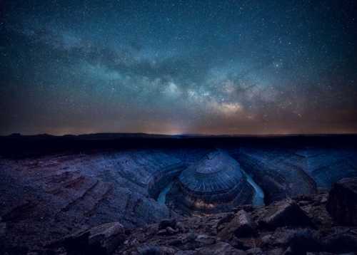 A stunning night sky filled with stars above a deep, rocky canyon landscape, illuminated by the Milky Way.