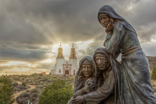 A bronze statue of three women in prayer, with a church and dramatic clouds in the background.
