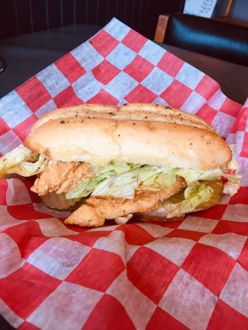 A sandwich with fried fish, lettuce, and pickles on a toasted bun, served on red and white checkered paper.