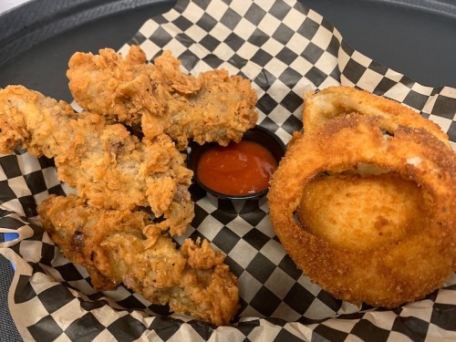 A tray with fried chicken tenders, a side of dipping sauce, and a crispy onion ring on a checkered paper.
