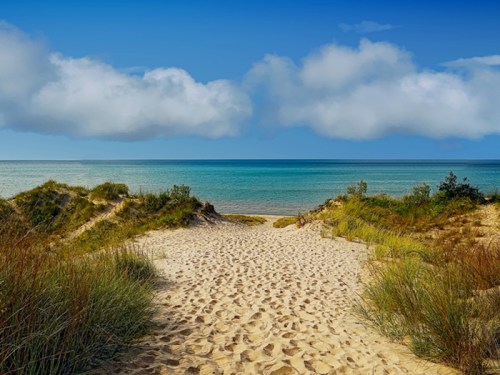 A sandy path leads to a tranquil beach with gentle waves and a blue sky dotted with clouds.