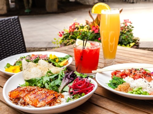 A table with three colorful dishes: salmon with rice, a salad, and a vibrant drink, surrounded by flowers.
