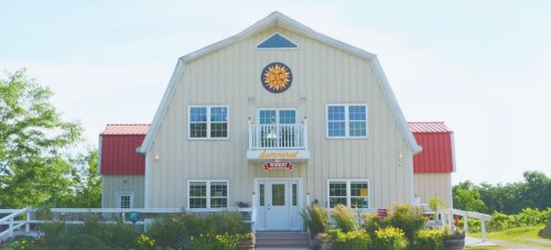 A large, light-colored barn-style building with a red roof and sun emblem, surrounded by greenery.