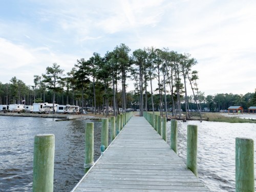 A wooden dock extends into a calm lake, surrounded by trees and RVs in the background under a clear sky.