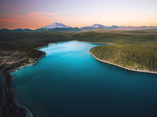 Aerial view of a serene lake surrounded by mountains and dense forest under a colorful sunset sky.