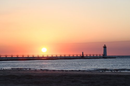 A serene sunset over the ocean, with a lighthouse silhouetted against the colorful sky and a pier extending into the water.