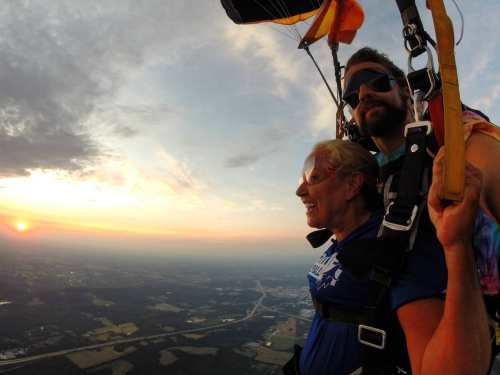 A woman smiles while tandem skydiving, with a sunset and landscape visible below.