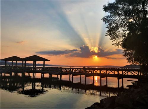 Sunset over a calm lake, with rays of light shining through clouds and a wooden dock silhouetted in the foreground.