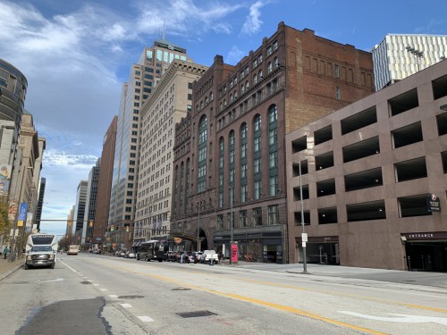 A city street lined with tall buildings, including historic and modern architecture, under a partly cloudy sky.