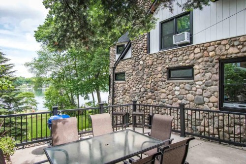 A stone house with large windows overlooks a patio with a table and chairs, surrounded by trees and a lake view.