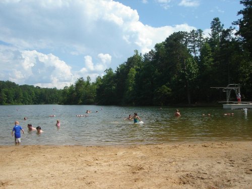 A sandy beach by a lake with people swimming and relaxing under a blue sky with clouds and trees in the background.