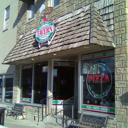 Exterior of a tavern with a stone facade, featuring large windows displaying the menu: pizza, pasta, and chicken.
