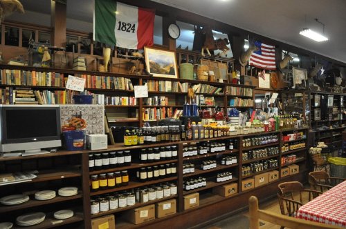 A cozy store interior filled with books, jars of preserves, and vintage decor, featuring flags and rustic shelves.