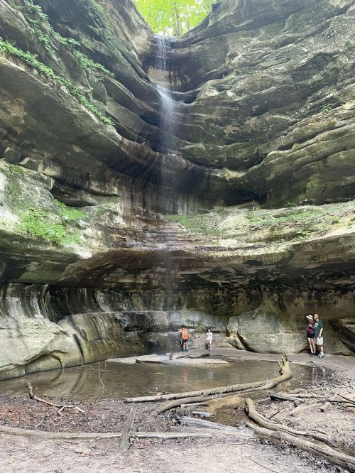 A waterfall cascades down rocky cliffs into a small pool, with people exploring the area beneath the natural formation.