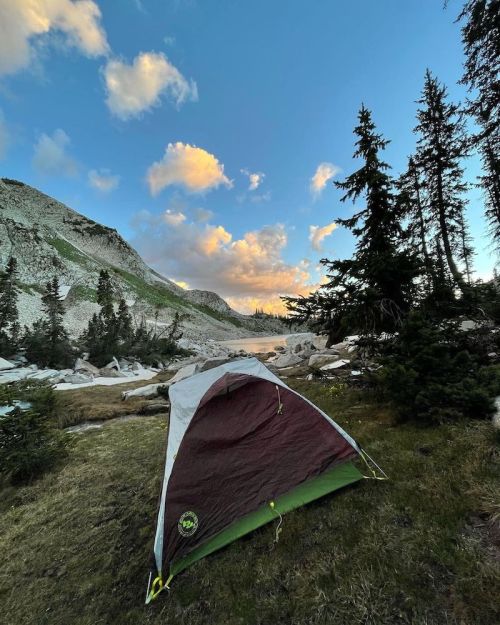 A tent set up in a scenic mountain landscape with trees, rocks, and a blue sky dotted with clouds.